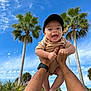 baby, child, person, hands, cap, smiling, outdoor, sky, blue_sky, clouds, palm_tree, greenery, nature, summer, sunny, happy, portrait, casual_clothing, barefoot, playful