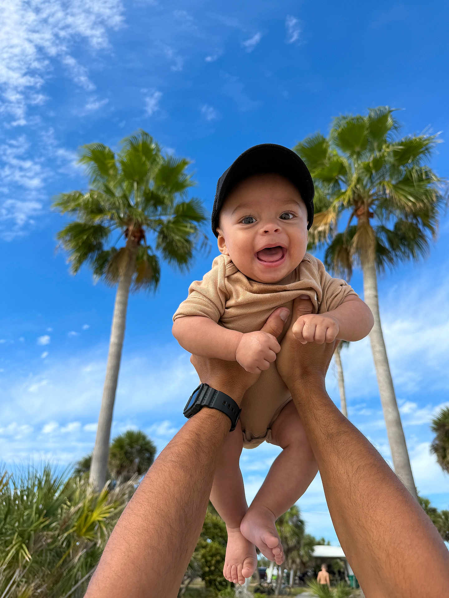 Roman is registered to the contest to win money with this photo: baby, child, person, hands, cap, smiling, outdoor, sky, blue_sky, clouds, palm_tree, greenery, nature, summer, sunny, happy, portrait, casual_clothing, barefoot, playful