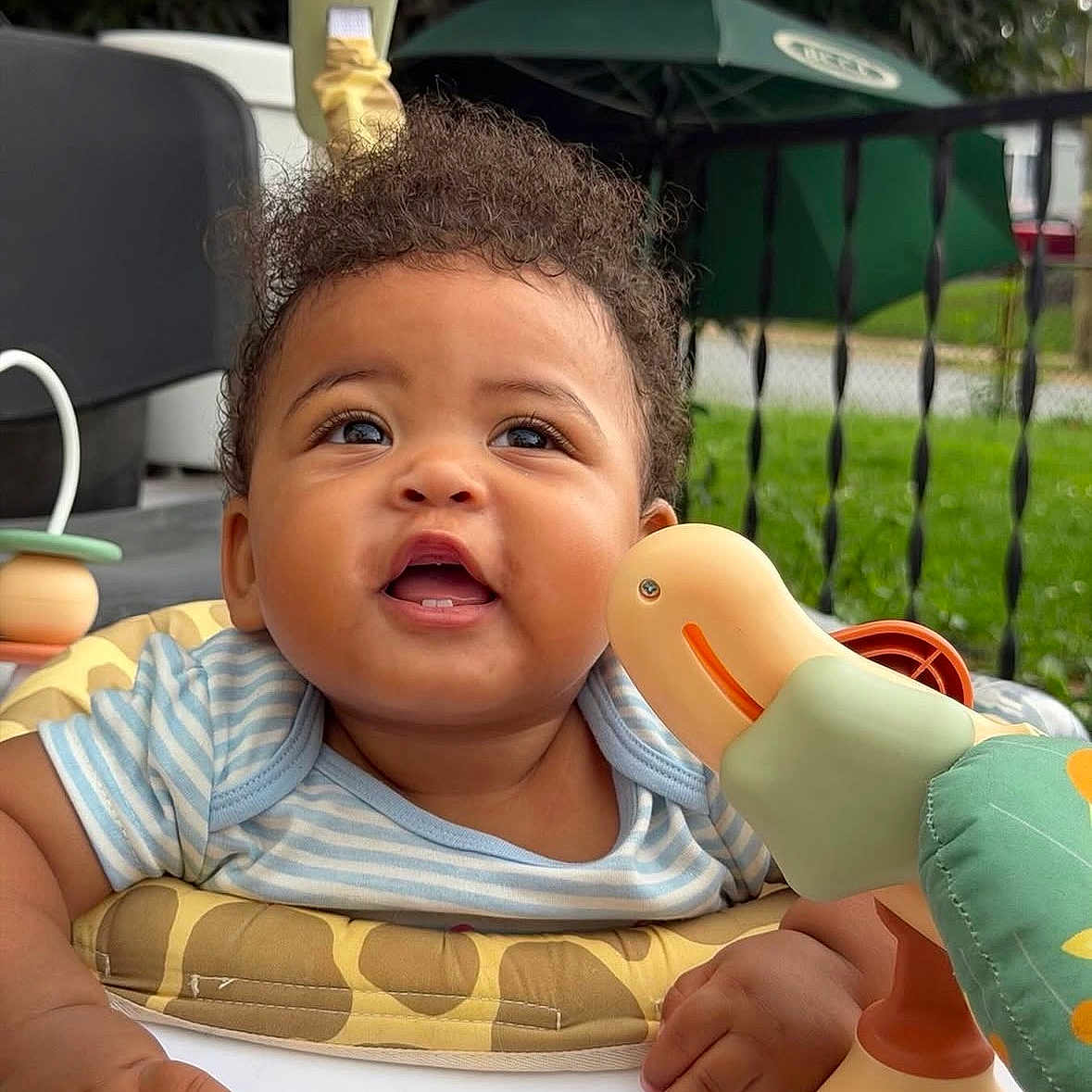 Layla is registered to the contest to win money with this photo: baby, child, curly_hair, toy, duck, smiling, outdoor, play, infant, hands, clothing, striped_shirt, face, happy, seat, greenery, umbrella, fence, closeup, portrait