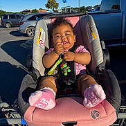 Layla is registered to the contest to win money with this photo: baby, car_seat, shopping_cart, parking_lot, toy, pink_socks, pink_shirt, child, sunlight, shadow, outdoor, vehicle, green_toy, happy, cute, infant, seatbelt, curly_hair, smiling, person