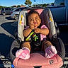 baby, car_seat, shopping_cart, parking_lot, toy, pink_socks, pink_shirt, child, sunlight, shadow, outdoor, vehicle, green_toy, happy, cute, infant, seatbelt, curly_hair, smiling, person