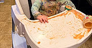 Wilder is registered to the contest to win money with this photo: baby, high_chair, food, mess, baby_food, smearing, clothing, face, floor, foot, kitchen, jar, table, plastic, child, expression, curious, indoor, person, seat