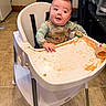baby, high_chair, food, mess, baby_food, smearing, clothing, face, floor, foot, kitchen, jar, table, plastic, child, expression, curious, indoor, person, seat
