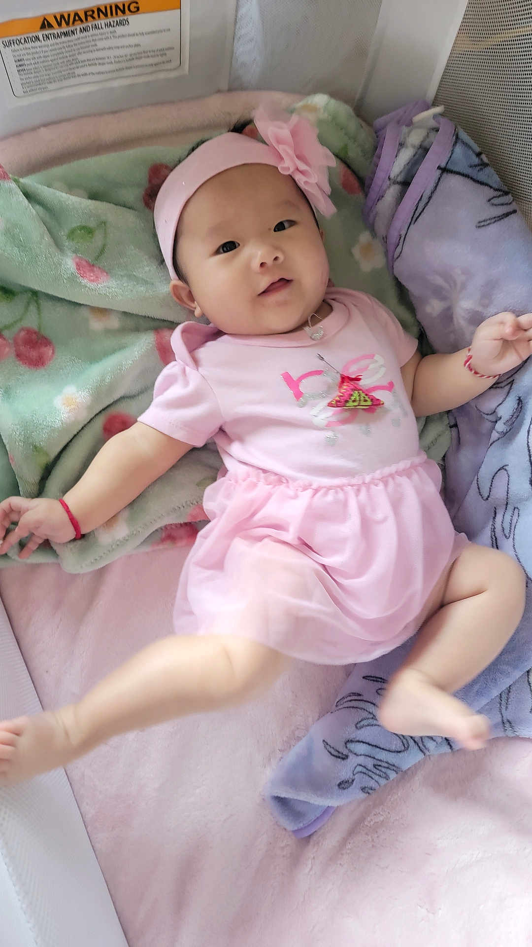 Lily is registered to the contest to win money with this photo: baby, infant, pink_dress, headband, flower, blanket, soft_texture, lying_down, smile, cute, child, indoors, cozy, pastel_colors, person, portrait, happy, young_child, sleeping_area, toddler
