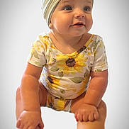 Paisley is registered to the contest to win money with this photo: baby, child, headband, bow, onesie, sunflower, floral, sitting, hands, feet, portrait, cute, blue_eyes, soft_background, indoor, infant, smiling, skin, clothing, person