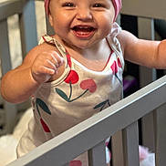 Paisley is registered to the contest to win money with this photo: baby, child, crib, headband, pink_bow, smile, happy, cute, infant, cherry_pattern, clothing, furniture, nursery, indoors, person, standing, hand, face, joy, playful