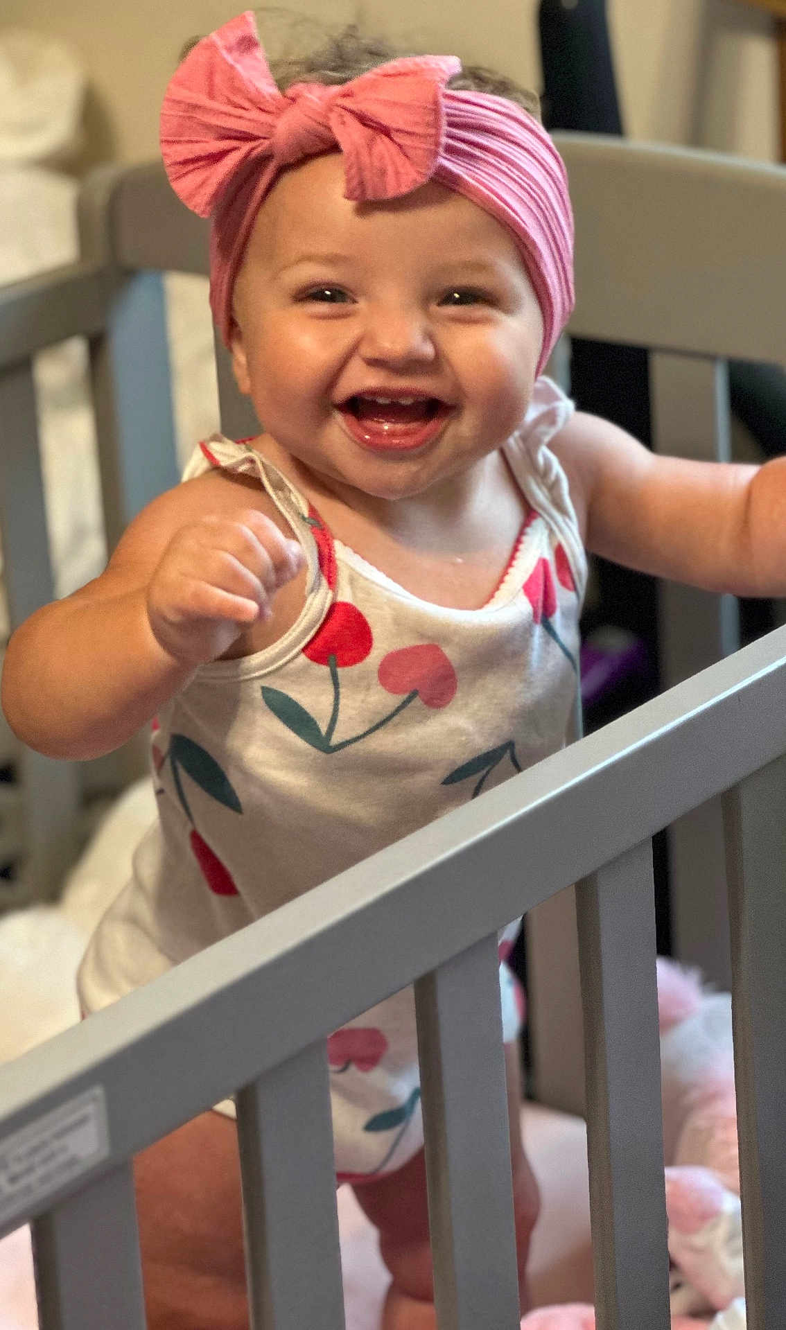 Paisley is registered to the contest to win money with this photo: baby, child, crib, headband, pink_bow, smile, happy, cute, infant, cherry_pattern, clothing, furniture, nursery, indoors, person, standing, hand, face, joy, playful