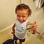 child, toddler, curly_hair, smiling, money, dollar_bill, white_shirt, graphic_tshirt, socks, tile_floor, bathroom, rug, door, towel, indoor, person, standing, cute, playful, expression