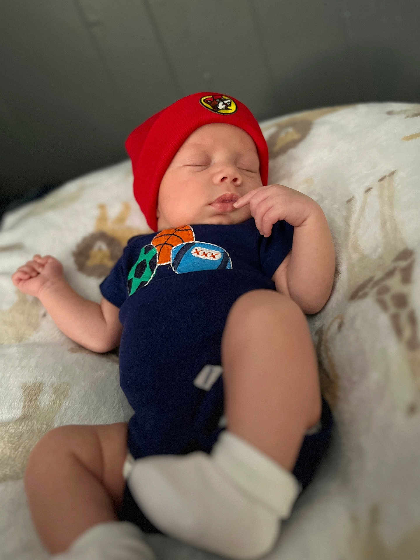 Kessiah is registered to the contest to win money with this photo: baby, sleeping, red_hat, onesie, sports_balls, blanket, infant, peaceful, cute, child, soft_focus, resting, clothing, newborn, indoor, person, portrait, headwear, sock, babywear
