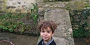 Zean participe au concours pour gagner de l'argent avec cette photo : child, boy, curly_hair, jacket, black_and_white, sneakers, stone_bridge, outdoor, smile, standing, stone_wall, door, grass, plants, water, casual_clothing, young_child, portrait, playful, nature