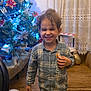 child, smiling, curly_hair, plaid_shirt, toy, christmas_tree, holiday_decorations, blue_lights, red_poinsettias, lace_curtains, wooden_floor, indoor, happy, festive, person, holiday, decor, balloon, furniture, home