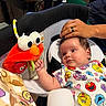 baby, infant, child, portrait, sitting, bouncer, elmo, plush_toy, sesame_street, colorful_clothing, onesie, wide_eyed, indoor, home, desk, computer, adult_hand, hand, toy, happy