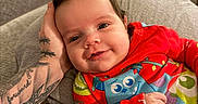 Saylor joined the competition — help win amazing prizes! baby, child, infant, smiling, portrait, close_up, indoors, couch, blanket, red_clothing, colorful_clothes, tattoo, arm, hand, face, eyes, happy, sitting, living_room, family