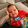 baby, child, infant, smiling, portrait, close_up, indoors, couch, blanket, red_clothing, colorful_clothes, tattoo, arm, hand, face, eyes, happy, sitting, living_room, family