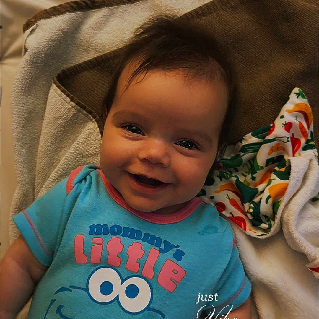 Saylor joined the competition — help win amazing prizes! baby, infant, child, smiling, portrait, lying_down, indoor, blanket, bedding, blue_shirt, cookie_monster, cartoon_character, colorful_clothing, cozy, happy, face, dark_hair, playful, text_overlay, closeup