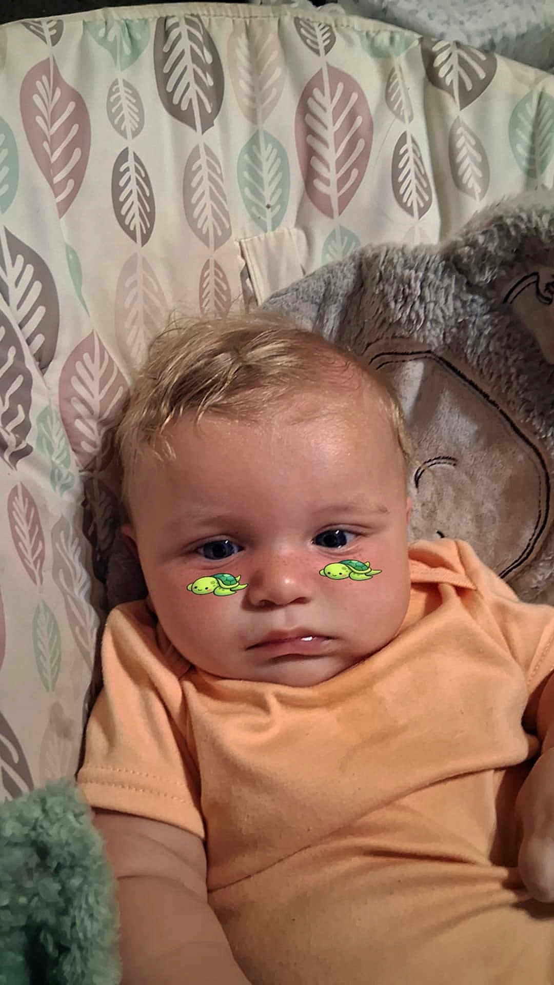 Theo is registered to the contest to win money with this photo: baby, infant, child, face, blue_eyes, peach_clothing, leaf_pattern, seat, blanket, turtle_stickers, cute, expression, skin, hair, indoor, soft_texture, comfort, portrait, young, closeup