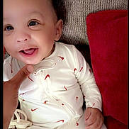 Remaje joined the competition — help win amazing prizes! baby, infant, smiling, onesie, white_clothing, red_pattern, couch, pillow, hand, face, happy, child, indoors, cute, portrait, young, person, seated, fabric, texture