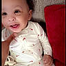 baby, infant, smiling, onesie, white_clothing, red_pattern, couch, pillow, hand, face, happy, child, indoors, cute, portrait, young, person, seated, fabric, texture