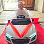 toddler, child, toy_car, audi_sport, red_ribbons, white_dress, headpiece, red_carpet, doorway, stone_floor, indoors, sunlight, serious_expression, vehicle, small_car, decorated_car, baby, portrait, person, floor