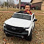 toddler, child, toy_car, white, outdoor, grass, smiling, happy, chevrolet, vehicle, playful, ball, curly_hair, small_car, parked_car, house, tree, dry_grass, daytime, fun