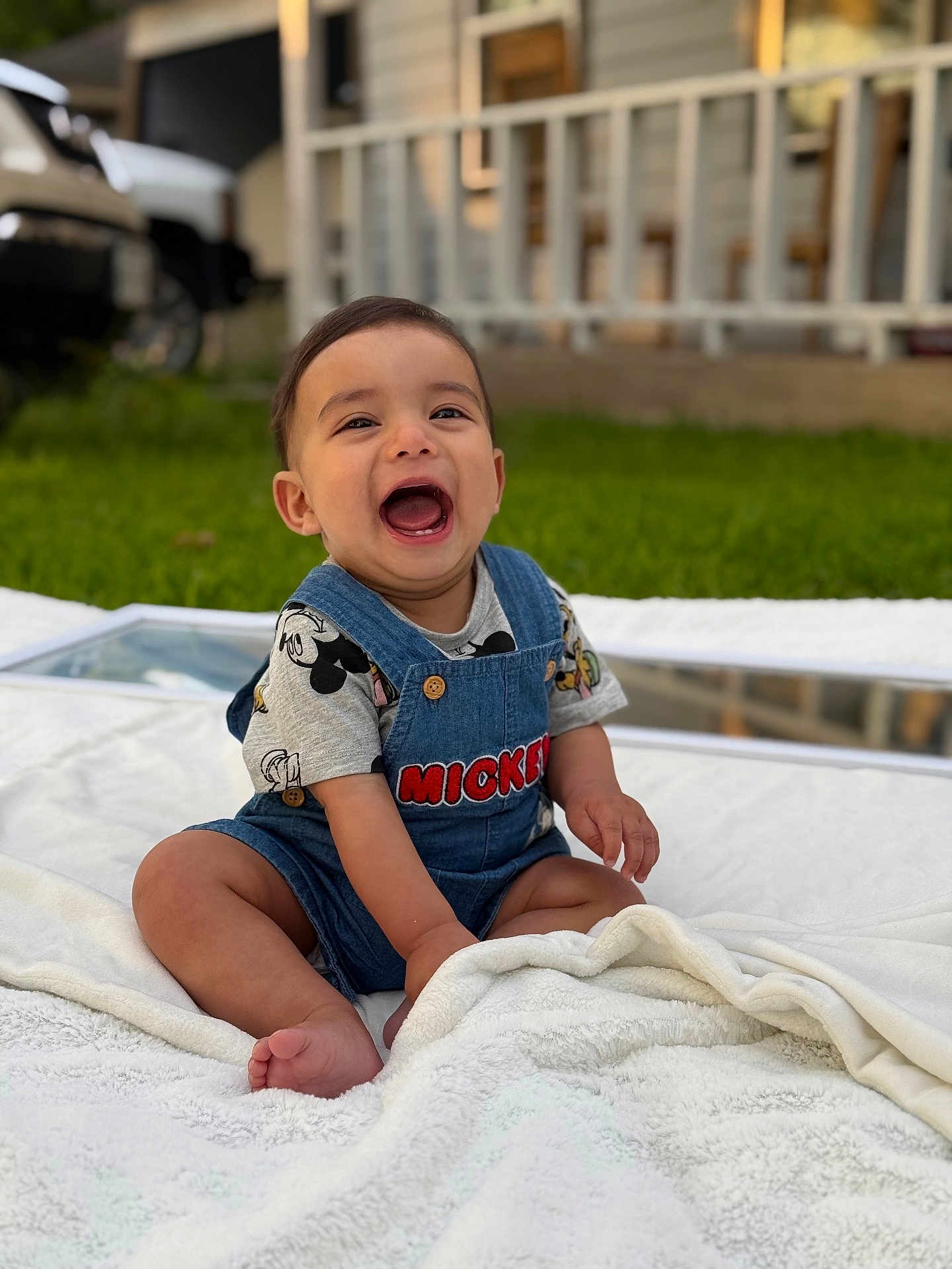 Eithan is registered to the contest to win money with this photo: baby, child, smiling, laughing, sitting, outdoor, blanket, denim_overall, mickey_mouse, shirt, grass, porch, vehicle, foot, hand, person, face, happy, clothing, daylight
