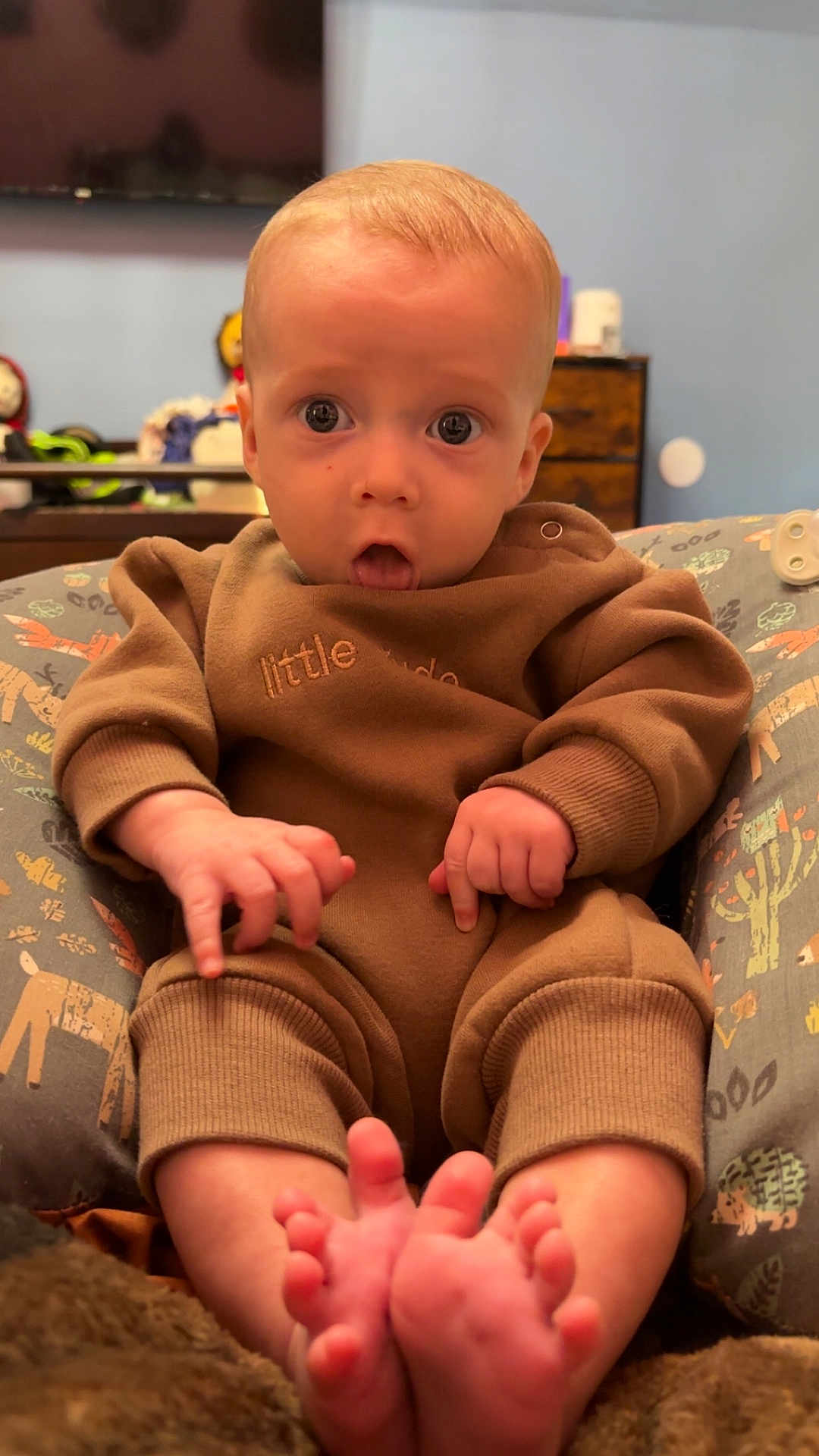 Bowen joined the competition — help win amazing prizes! baby, infant, child, face, eyes, hands, feet, toes, onesie, brown_clothing, support_pillow, sitting, surprised_expression, portrait, indoor, nursery, dresser, television, blanket, soft_toys