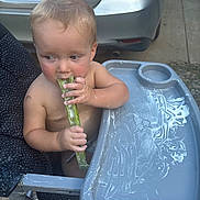 Bowen is registered to the contest to win money with this photo: baby, child, toddler, high_chair, cucumber, eating, snack, food, bare_chested, outdoor, driveway, car, parked_car, license_plate, infant, portrait, sitting, hands, blond_hair, side_eye