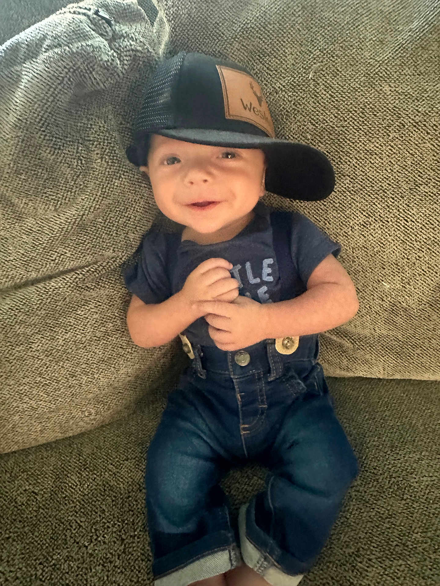 Wesley is registered to the contest to win money with this photo: baby, child, smiling, cap, hat, denim, overalls, couch, textile, clothing, person, infant, happy, portrait, casual, cute, sitting, indoor, face, young