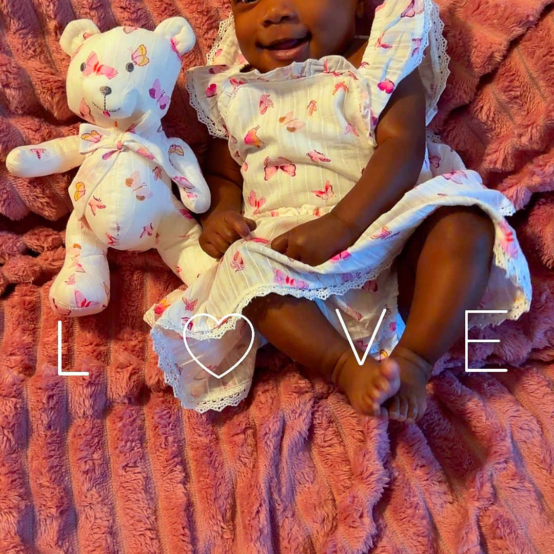 Victoria is registered to the contest to win money with this photo: baby, child, infant, smiling, portrait, sitting, lying_down, stuffed_bear, toy, blanket, pink, dress, headband, bow, heart, text_overlay, indoor, cuddly, happy, cute