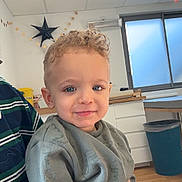Talha participe au concours pour gagner de l'argent avec cette photo : child, toddler, boy, curly_hair, blue_eyes, smile, sweatshirt, indoor, room, window, ceiling, star_decor, trash_can, wooden_floor, cabinet, adult_hand, portrait, happy, daylight, decorations