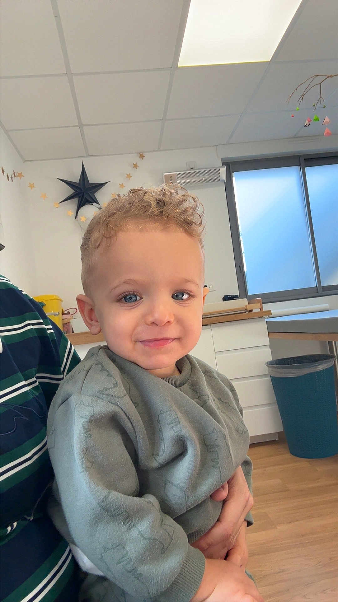 Talha participe au concours pour gagner de l'argent avec cette photo : child, toddler, boy, curly_hair, blue_eyes, smile, sweatshirt, indoor, room, window, ceiling, star_decor, trash_can, wooden_floor, cabinet, adult_hand, portrait, happy, daylight, decorations