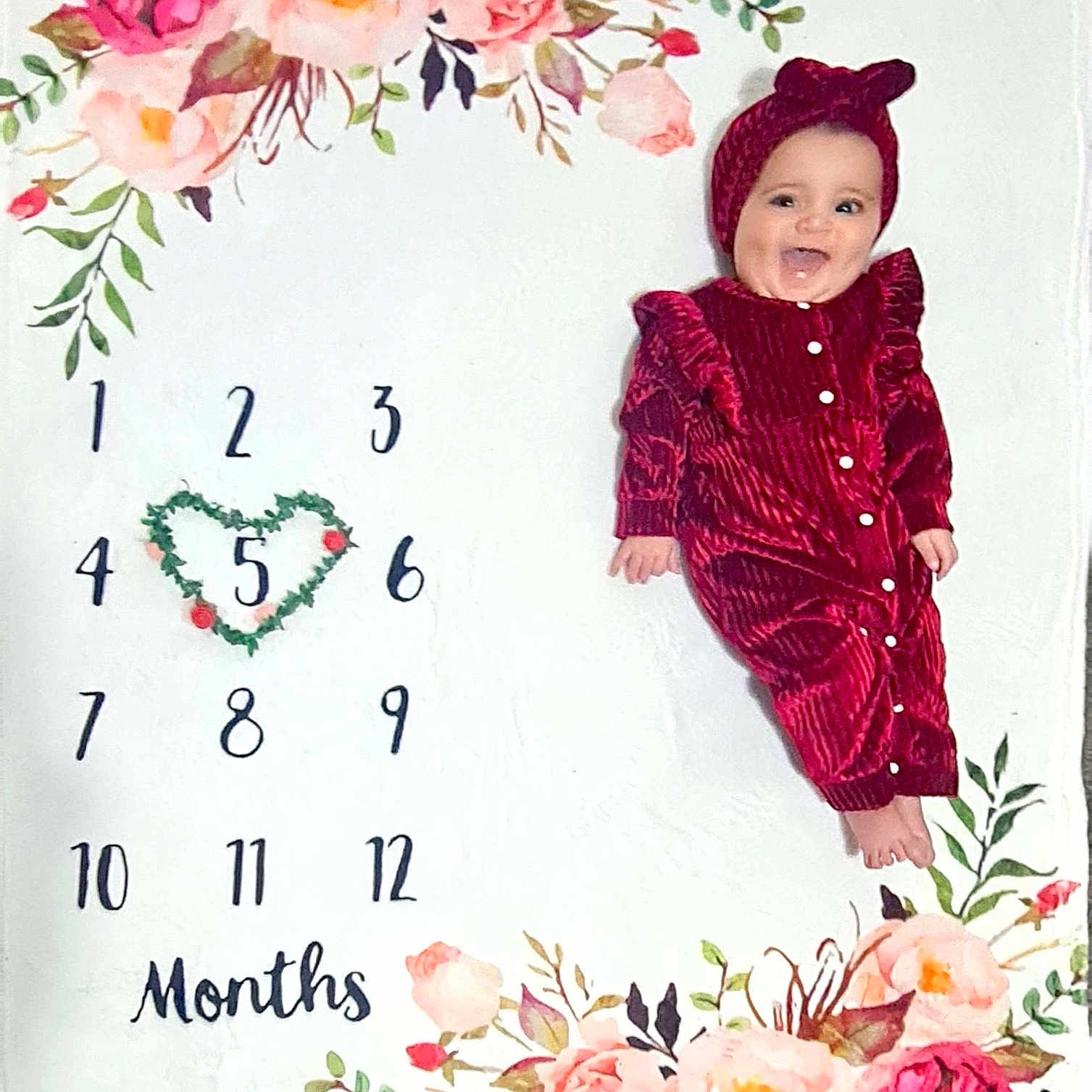 Camellia is registered to the contest to win money with this photo: baby, celebration, child, cute, floral_pattern, greenery, happy, headband, heart_shape, indoors, infant, lying_down, milestone_blanket, months, numbers, portrait, red_clothing, smiling, white_background, young_child