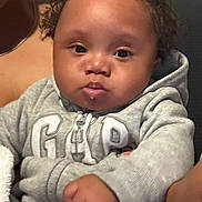 Noah is registered to the contest to win money with this photo: baby, child, curly_hair, hoodie, gray_clothing, indoor, couch, blanket, face, person, infant, portrait, fist, cute, expression, young_child, seated, home, soft_lighting, casual_wear