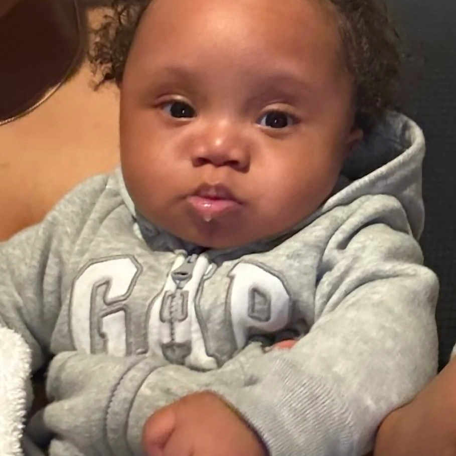 Noah is registered to the contest to win money with this photo: baby, blanket, casual_wear, child, couch, curly_hair, cute, expression, face, fist, gray_clothing, home, hoodie, indoor, infant, person, portrait, seated, soft_lighting, young_child