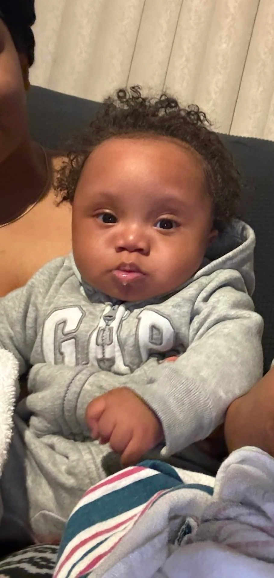 Noah is registered to the contest to win money with this photo: baby, child, curly_hair, hoodie, gray_clothing, indoor, couch, blanket, face, person, infant, portrait, fist, cute, expression, young_child, seated, home, soft_lighting, casual_wear