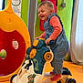 Giulia participe au concours pour gagner de l'argent avec cette photo : toddler, child, toy_vehicle, indoor_play_area, blue_romper, orange_shirt, smiling, happy, playground_equipment, colorful, plastic_toy, yellow, red, green_wall, curtain, floor_mats, feet, socks, young_child, playtime