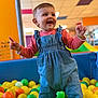 toddler, child, ball_pit, indoor, play_area, colorful, smiling, denim, romper, pink_shirt, yellow_balls, green_balls, orange_balls, happy, cute, playful, young_child, bright, fun, person