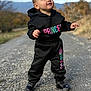 toddler, child, outdoor, path, hoodie, pants, black_clothing, colorful_text, shoes, standing, curious, nature, gravel, young_child, casual_wear, portrait, daylight, expression, small_person, street