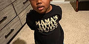 Harlem is registered to the contest to win money with this photo: child, toddler, indoor, carpet, dresser, table, shadow, black_shirt, text_on_clothing, jeans, sneakers, red_shoes, standing, looking_up, face, person, casual_clothing, home, furniture, curly_hair