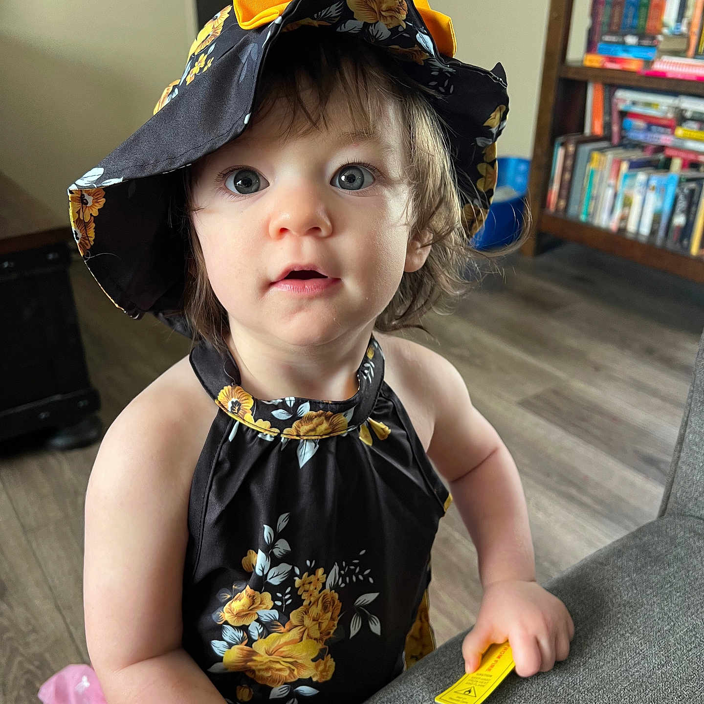 Vyolett is registered to the contest to win money with this photo: black, blue_eyes, bookshelf, casual, child, clothing, curious, expression, floor, floral_pattern, furniture, hand, hat, indoor, person, portrait, toddler, wooden_floor, yellow, young_child
