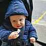 baby, child, blue_jacket, hood, stroller, food, hand, face, outdoor, sidewalk, road, curious, winter_clothing, young_child, cute, person, seatbelt, urban, expression, walking_path