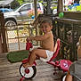 child, toddler, tricycle, diaper, porch, wooden_floor, outdoor, smiling, play, toy, vehicle, greenery, flower_pot, truck, fence, sunlight, decorations, happy, person, summer
