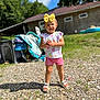 casual_clothing, child, daylight, grass, gravel, greenery, happy, house, nature, outdoor, person, playful, sandals, shorts, smile, standing, summer, sunny, toddler, yellow_bow