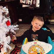Weston is registered to the contest to win money with this photo: child, smiling, toy_car, christmas_tree, ornament, indoor, holiday, blanket, carpet, quilt, hoodie, happy, furniture, decor, toy, face, person, car, play, festive