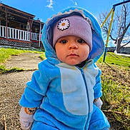 Malakai is registered to the contest to win money with this photo: toddler, child, blue_onesie, purple_hat, daisy_patch, outdoor, grass, sky, clouds, porch, sidewalk, gloves, winter_clothing, playground, swing, trees, sunny, face, curious, seated