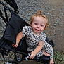 toddler, child, stroller, curly_hair, blue_eyes, leopard_print, outdoor, gravel, dirt, grass, footwear, hand, smile, person, seat, black, clothing, baby, cute, portrait