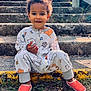 child, toddler, stairs, outdoor, sunlight, onesie, patterned_clothing, smiling, curly_hair, red_shoes, grass, wooden_railing, nature, portrait, happy, casual, sitting, daylight, concrete, playful