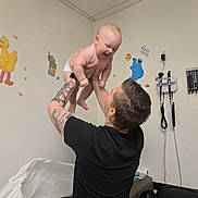 Creed Taylor is registered to the contest to win money with this photo: baby, man, tattoos, black_shirt, diaper, pediatric_office, medical_equipment, wall_decals, sesame_street, big_bird, cookie_monster, room, ceiling, lamp, chair, table, paper_sheet, joy, playful, bonding