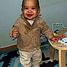 Elaïjah a rejoint le concours — aidez-le/la à gagner de superbes lots ! beige_pants, brown_jacket, child, expression, face, floor_mat, furniture, hand, happy, indoors, person, play_mat, smiling, standing, toddler, toy, wall, white_shoes, wooden_table, young_child