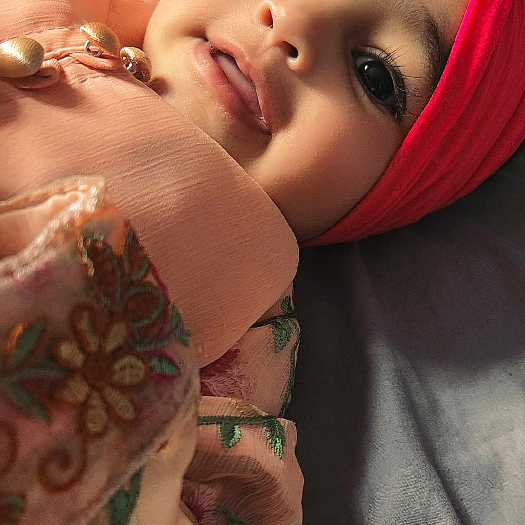 Nyra is registered to the contest to win money with this photo: baby, child, close_up, curious_eyes, cute, embroidery, eyelashes, fabric, face, happy, head_wrap, indoors, infant, lying_down, peach_clothing, pink, portrait, smile, soft_lighting, texture